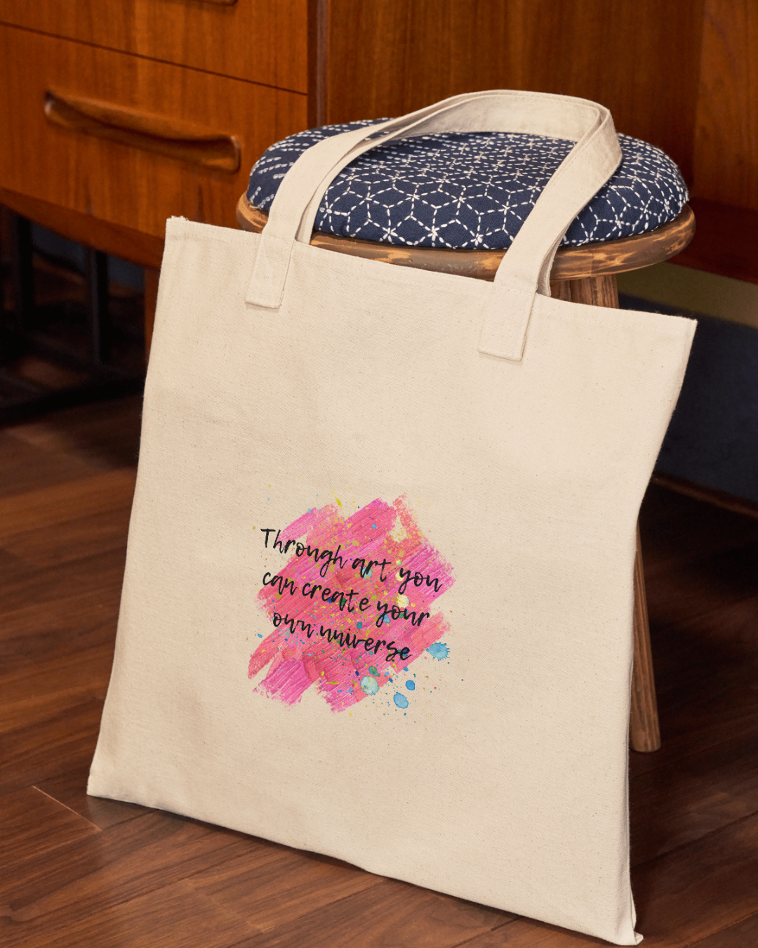 Sacoșa tip tote bag din bumbac organic sprijinită de un taburet din lemn. Sacosa este cu imprimeu ce imita urmele de vopsea in nuanțe prediminante de roz cu stropi de vopsea diverse culori și cu citatul ”Through art you can create your own universe” scris cu text negru cu contur alb intr-un font ce imită o tușă de pensulă