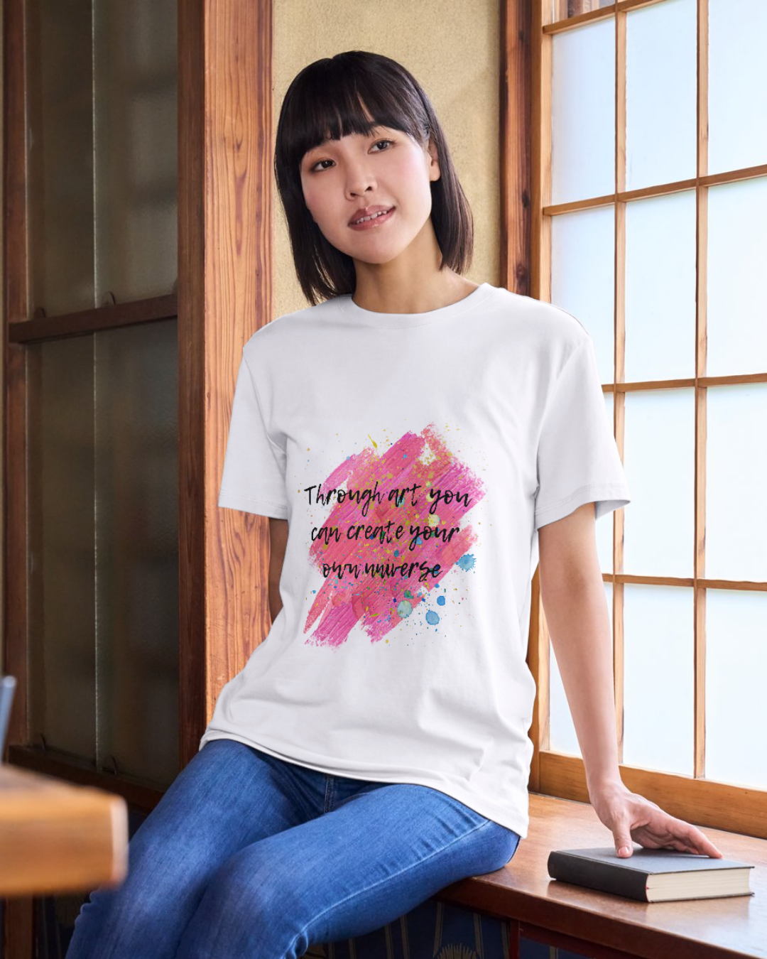 Modelul feminin care sta sprijinit
de un pervaz într-o bibliotecă, poartă un tricou unisex alb din bumbac organic cu imprimeu ce imita urmele de vopsea in nuanțe prediminante de roz cu stropi de vopsea diverse culori și cu citatul ”Through art you can create your own universe” scris cu text negru cu contur alb intr-un font ce imită o tușă de pensulă