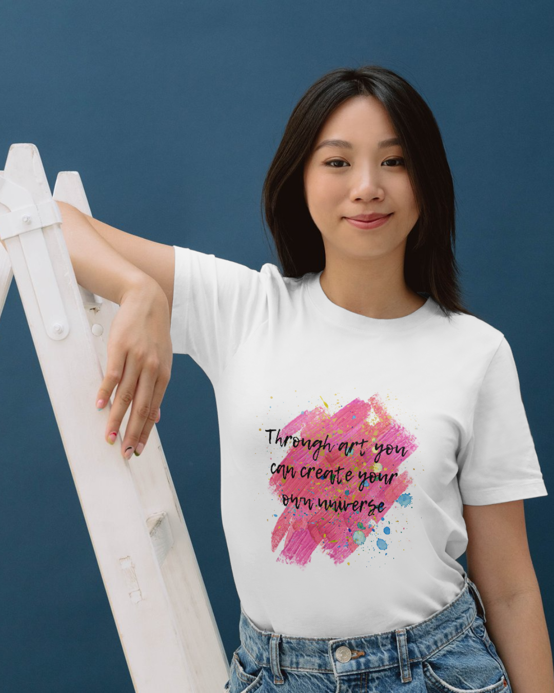 Modelul feminin care sta sprijinit
de o scară poartă un tricou unisex alb din bumbac organic cu imprimeu ce imita urmele de vopsea in nuanțe prediminante de roz cu stropi de vopsea diverse culori și cu citatul ”Through art you can create your own universe” scris cu text negru cu contur alb intr-un font ce imită o tușă de pensulă