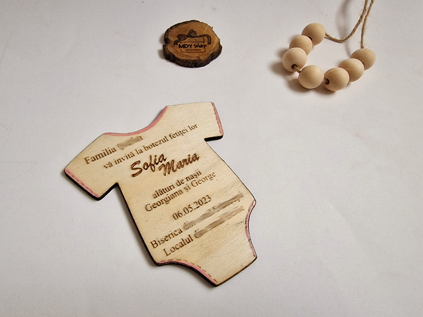 invitație pentru botez din placaj de lemn de plop, tăiat laser în formă de body pentru bebeluși și cu textul gravat. Pe invitație scrie ”Familia...vă invită la botezul fetiței lor Sofia Maria alături de nașii Georgiana & George 06.05.2023 Biserica... Localul...”. În partea de sus a imagini este o secțiune de creangă pe care este gravat logo-ul MDY Shop și lângă este o sfoară cu mărgele de lemn