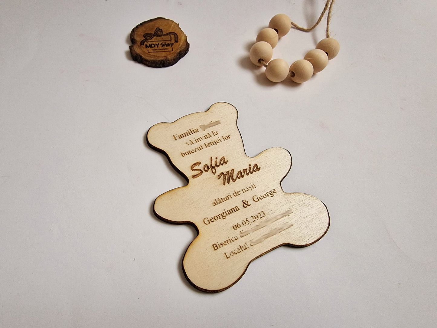invitație pentru botez din placaj de lemn de plop, tăiat laser în formă de ursuleț de pluș și cu textul gravat. Pe invitație scrie ”Familia...vă invită la botezul fetiței lor Sofia Maria alături de nașii Georgiana & George 06.05.2023 Biserica... Localul...”. În partea de sus a imagini este o secțiune de creangă pe care este gravat logo-ul MDY Shop și lângă este o sfoară cu mărgele de lemn