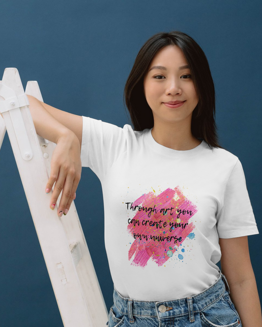 Modelul feminin care sta sprijinit 
 de o scară poartă un tricou unisex alb din bumbac organic cu imprimeu ce imita urmele de vopsea in nuanțe prediminante de roz cu stropi de vopsea diverse culori și cu citatul ”Through art you can create your own universe” scris cu text negru cu contur alb intr-un font ce imită o tușă de pensulă